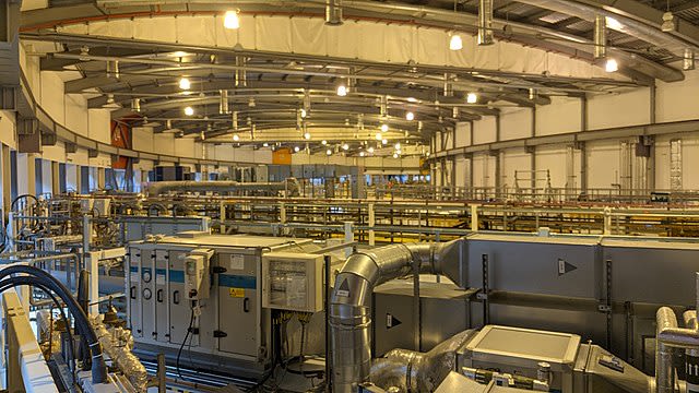 A picture taken from inside a synchrotron facility, in an enormous rooms that curves around in the distance, with large metal pipes all around and many cables linking pieces of equipment